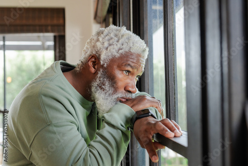 Senior African American man in sweatshirt gazing outside through window at home wearing smartwatch