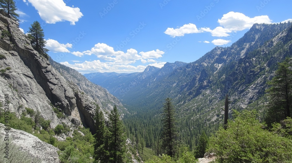 Fototapeta premium Majestic mountain valley scene with towering rock faces, lush green forests, and a vibrant blue sky dotted with fluffy white clouds. A breathtaking vista of nature's grandeur
