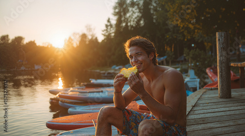 Young man enjoying a fruit snack on a wooden dock by the lake at sunset, with paddleboards lined up along the water's edge.