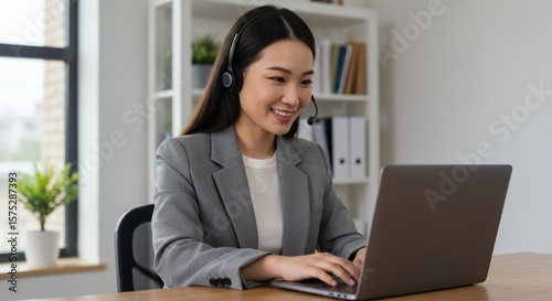 A smiling Asian woman in a grey blazer uses a headset and laptop at a desk in a bright office