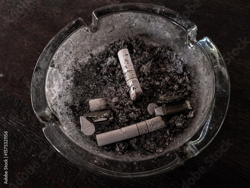 A used glass ashtray contains several extinguished cigarette butts and a pile of gray ash