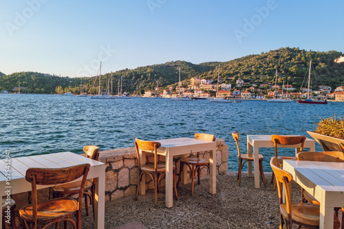Fototapeta Naklejka Na Ścianę i Meble -  Restaurant tables overlooking the bay of Vathy, Ithaca at sunset, with traditional colorful Ionian houses in the background, Ithaca, Greece