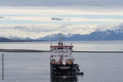 Punta Arenas, Chile - 12 June 2025: Aerial view of a sturdy ship slicing through the calm, steely waters, framed against a backdrop of snow-capped mountains under a hazy sky.
