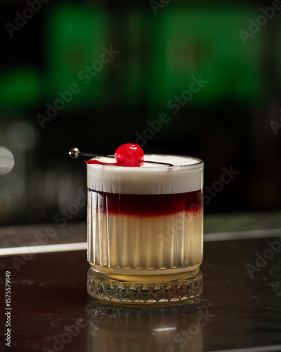 A striking layered cocktail features a rich red top over a pale yellow base, crowned with a thick white foam and garnished with a single vibrant red cherry on a skewer