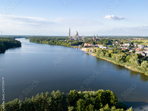 Aerial view of the Basilica of Our Lady of Lichen surrounded by lush forest, lake, and rural landscape under clear blue summer sky. Shot in Poland