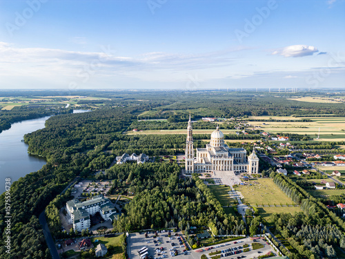 Aerial view of the Basilica of Our Lady of Lichen surrounded by lush forest, lake, and rural landscape under clear blue summer sky. Shot in Poland