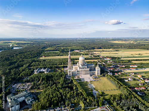 Aerial view of the Basilica of Our Lady of Lichen surrounded by lush forest, lake, and rural landscape under clear blue summer sky. Shot in Poland