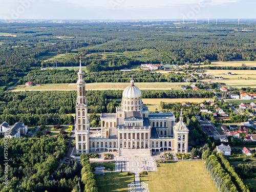 Aerial view of the Basilica of Our Lady of Lichen surrounded by lush forest, lake, and rural landscape under clear blue summer sky. Shot in Poland