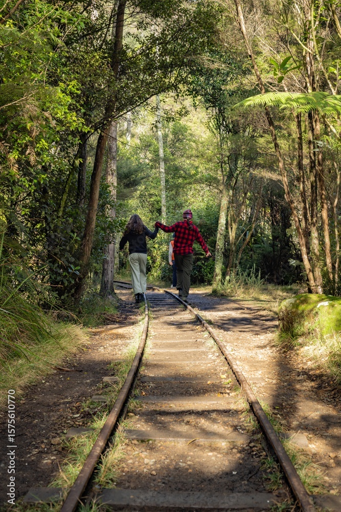 Obraz premium A couple holds hands while walking on an old railway track through a lush forest in New Zealand. They are enjoying a hike and exploring the natural beauty of the area. Karangahake Gorge, NZ