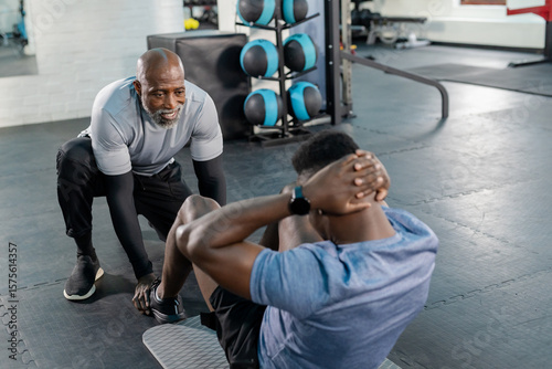 African American trainer bracing feet on mat, client performing situps at gym with ball rack
