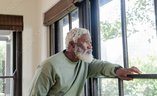 Senior African American man in sweatshirt leaning on window sill with blinds at home gazing outside