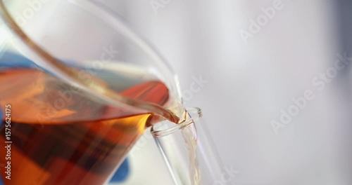 Stream of dark amber liquid reagent pours from large glass beaker into glass test tube. Focus on precise chemical transfer in laboratory setting