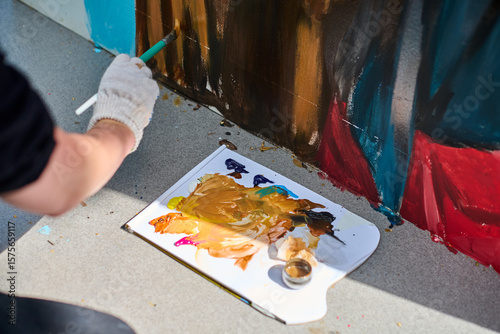 Focused view of adults hand painting mural outdoors. Gloved hand holds brush applying vivid red and blue hues on rough surface with precision and artistry