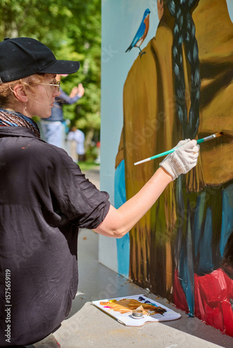 Young adult female artist with gloves and cap painting vibrant mural. Focused, intent expression. Sunlit park backdrop with greenery enhances creativity
