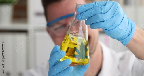 Scientist in gloves and protective eyewear holds conical flask with yellow liquid. Cannabis leaf indicates cannabis-based oil research in laboratory