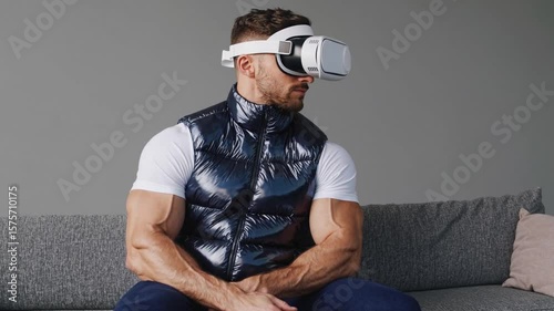Muscular man on grey sofa wearing VR headset in shiny navy puffer vest