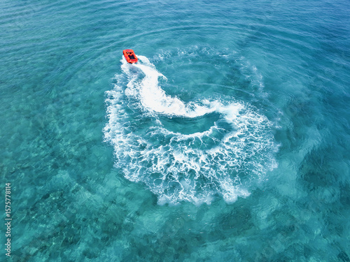 Aerial view of a vibrant orange boat carving swirling patterns in the turquoise waters, leaving a frothy white wake behind, Gulhi, Kaafu Atoll, Maldives.
