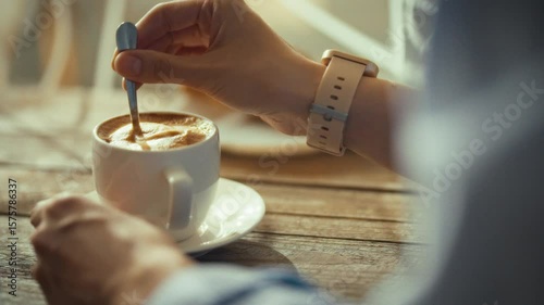 A person gently stirs a cup of coffee using a spoon while sitting at a table. The warm light of a sunny morning fills the cozy cafe, creating a peaceful atmosphere