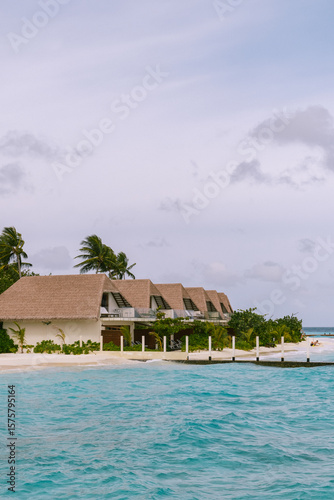 Fototapeta Naklejka Na Ścianę i Meble -  Tropical seaside resort with rows of overwater bungalows and palm trees swaying gently under a cloudy sky. A peaceful vacation setting by the calm turquoise ocean, perfect for relaxation and escape.
