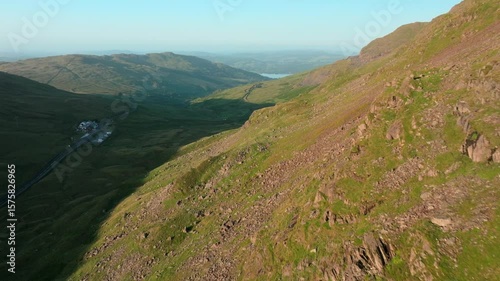 Flying close to mountainside illuminated by early morning sunshine. With mountain pass road snaking towards distant lake Windermere. Summer. Kirkstone Pass, Lake District, Cumbria, UK.