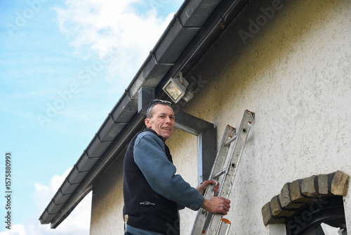 Professional electrician installing powerful automatic LED light with motion sensor under house roof, standing on ladder with outside of residential building, looking at camera