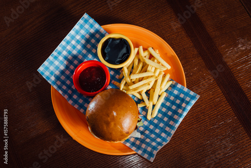 Top view of Bavarian-style beef burger with fries and sauce on a green plate over a blue checkered Bavarian napkin. Flat lay shot of traditional German-inspired comfort food.