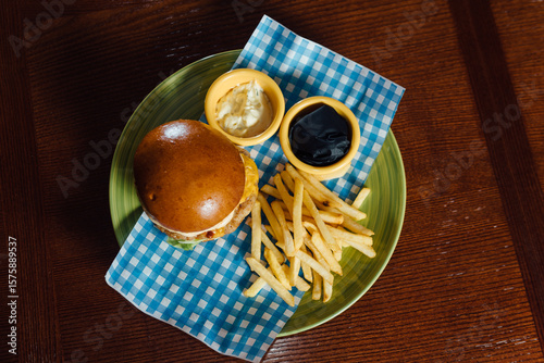 Top view of Bavarian-style chicken burger with fries and sauces on a green plate over a blue checkered Bavarian napkin. Flat lay shot of traditional October fest food.