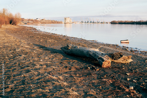Driftwood on quiet riverbank at sunset, industrial port visible in the distance