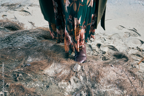 Woman in floral dress and boots standing on sandy beach with dry grass