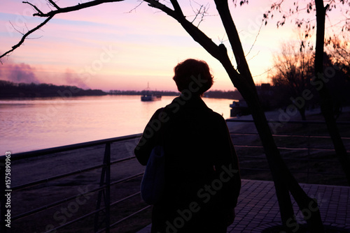 Silhouette of a woman watching sunset by river, tranquil evening urban scene