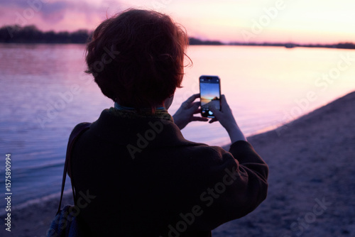 Woman photographing sunset by the river with smartphone on a tranquil evening
