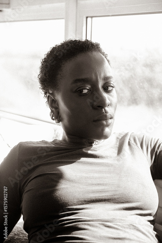 Portrait of a Black queer woman, sitting on the sofa looking to the side.