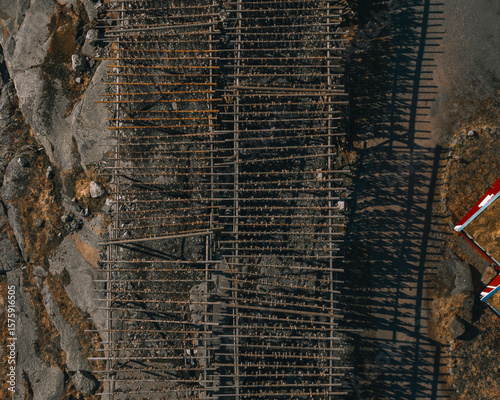 Aerial view of wooden racks stretching across rocky terrain, casting long shadows, near a building with a red-and-white roof, Reine, Nordland, Norway.