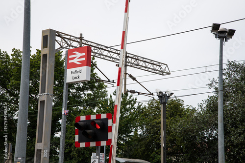 View of Enfield Lock railway station signage with level crossing barriers, overhead power lines, and CCTV—capturing everyday UK rail infrastructure in detail.