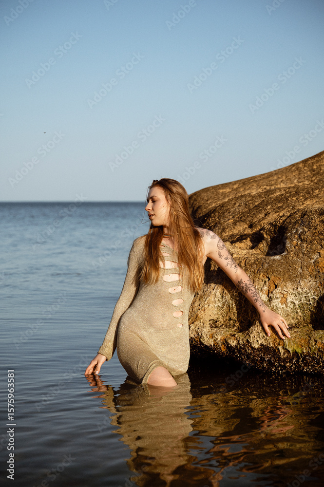 Fototapeta premium Blonde with long hair in sexy dress stands in sea against rock background.