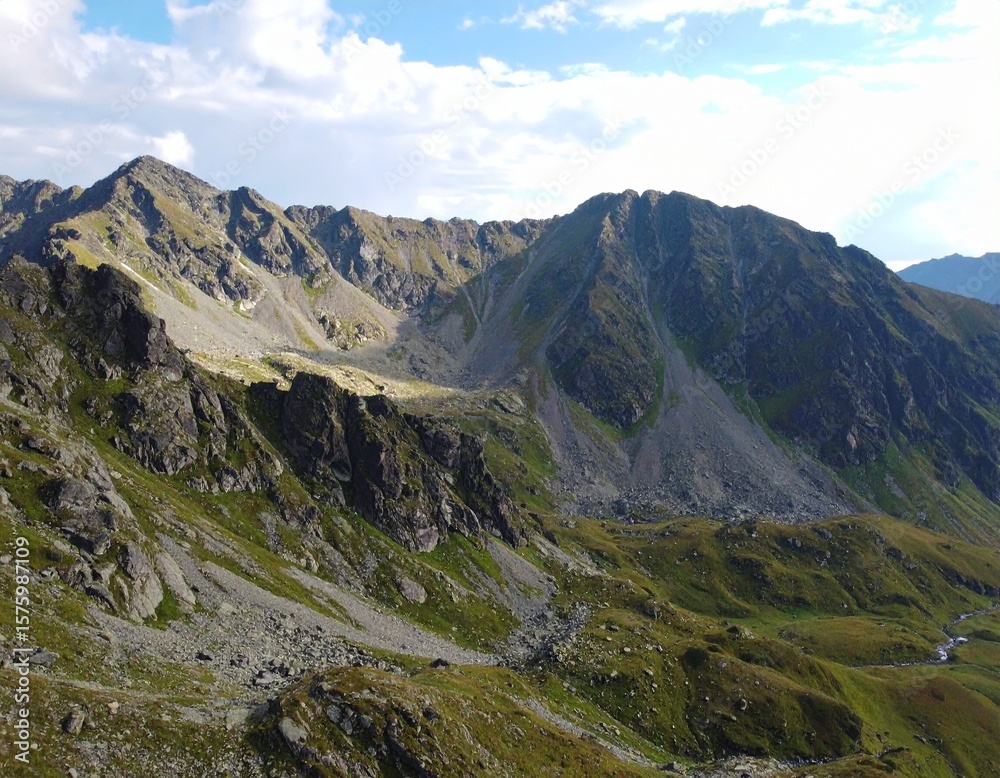 Fototapeta premium A mountainous landscape featuring rocky peaks and green slopes under a partly cloudy blue sky view from above