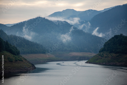 Misty morning over mountain lake in Retezat National Park. Ethereal fog drifting through forested Carpathian valleys creating atmospheric landscape