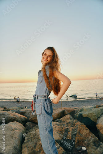 lifestyle woman in modern boho-western denim street style smiles happily outdoors by the rocky beach at sunset in film color warm tones