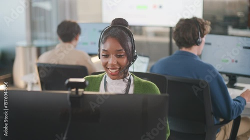 Confident African American female call center agent communicates with clients using headset and computer. She offers professional tech support at a modern office desk, smiling diverse office