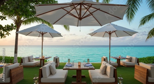 Tranquil beachfront dining area with three white umbrellas shading low wooden tables and cushioned seating on a verdant lawn overlooking a serene turquoise ocean at sunset