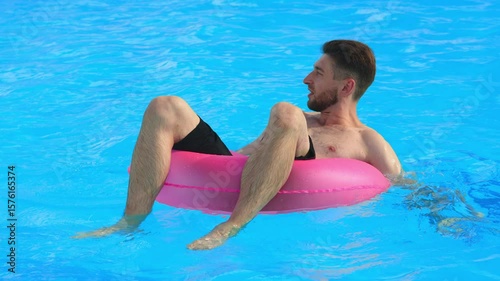 Young man relaxing on a pink inflatable ring, floating in an outdoor pool. Concept leisure, summer vibe, carefree mood.