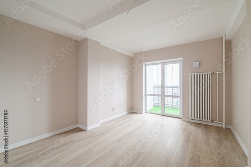 bright  empty room with light wood floors  beige walls  and a large window to a balcony. A white radiator stands against the wall