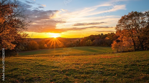 A vibrant autumn sunset casts golden light over a grassy field bordered by colorful fall trees under a partly cloudy sky.