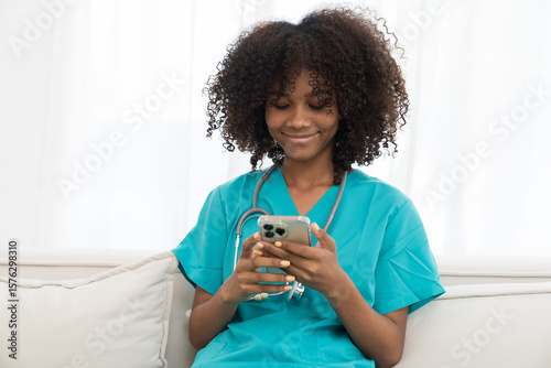 Female doctor sitting on sofa and using mobile phone in hospital. Female nurse wearing scrubs uniform in office at hospital