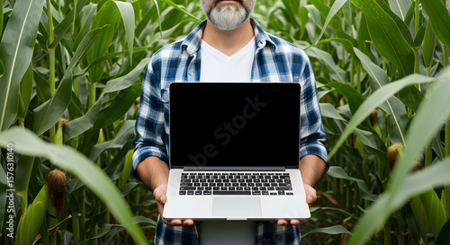 A farmer holds a laptop, standing amidst a lush cornfield, showcasing agricultural technology.