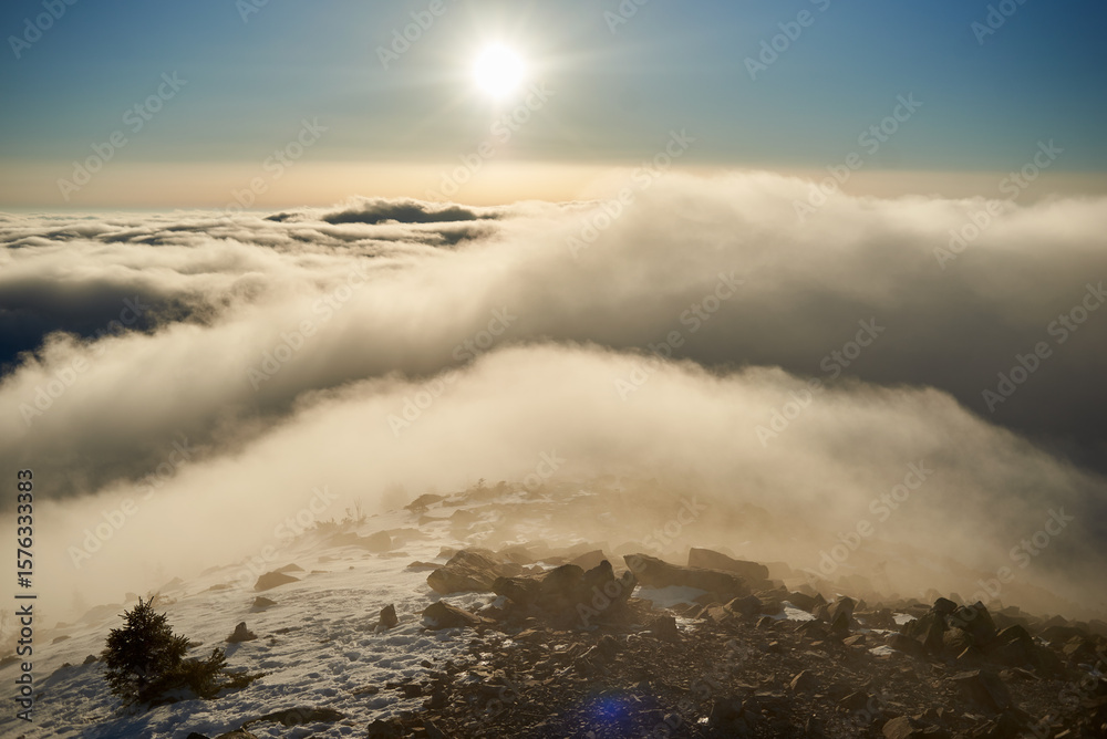 Obraz premium Sunrise over snow-covered mountain peak. Warm golden light of rising sun bathes clouds in soft hues of orange and pink. Snow and scattered rocks in foreground add texture and depth to scene.