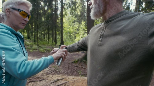 Mature Caucasian woman in trekking sunglasses spraying mosquito repellent on her husband protecting from insects while hiking together in summer forest on sunny day