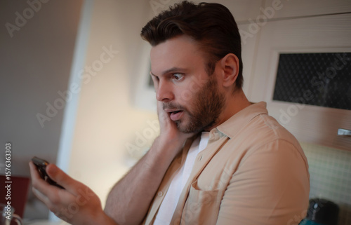 Surprised young man in casual clothes holds smartphone in a kitchen, looking shocked while reading unexpected news or message on his phone.