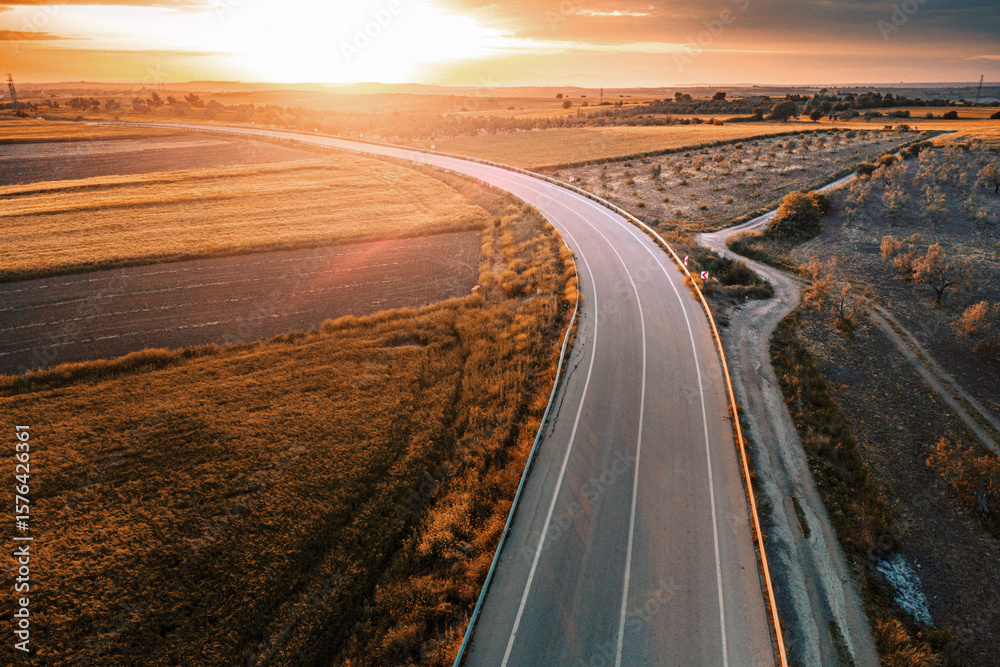 Fototapeta premium Aerial view of a winding road crossing colorful agricultural fields at sunset, creating a picturesque landscape