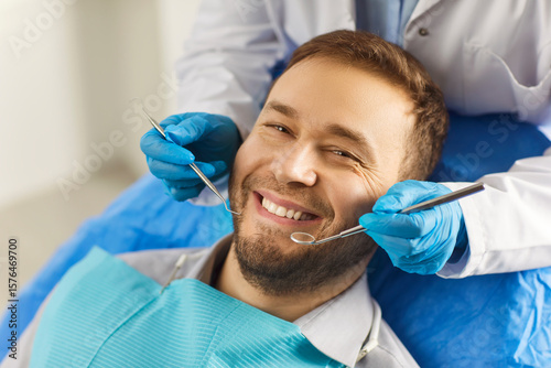 Dental health. Man sits comfortably in dentists chair, showing off white smile that exudes confidence. Dentist holds special tools in his hands for preventive and therapeutic examination of teeth.